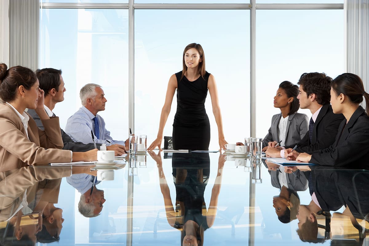 A woman in a black dress stands at the head of a boardroom glass table surrounded by other executives.