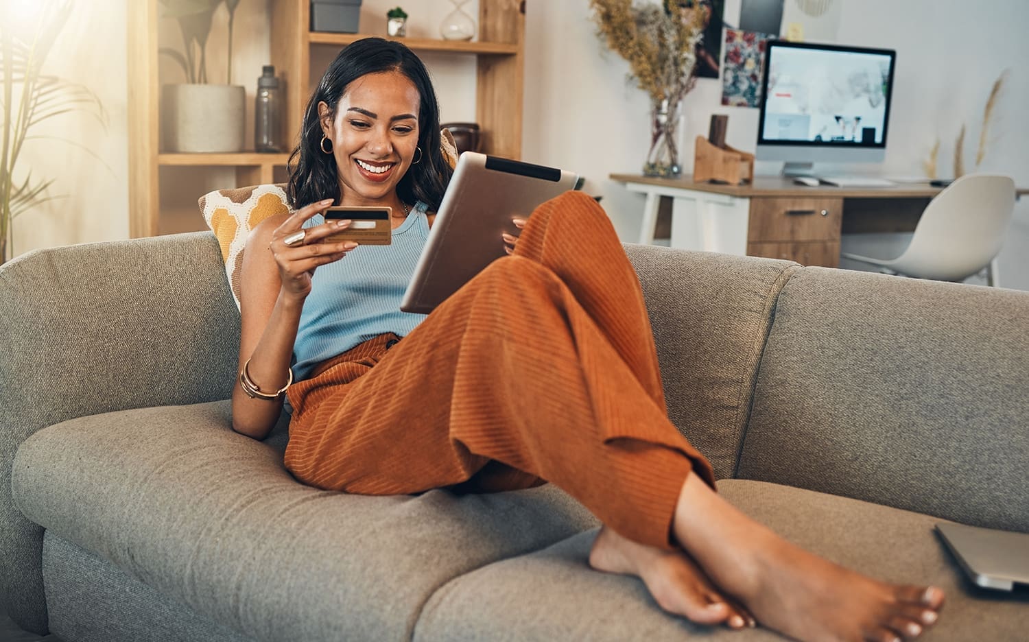 A woman reclines on her couch and smiles while looking at her debit card with a tablet in her lap.