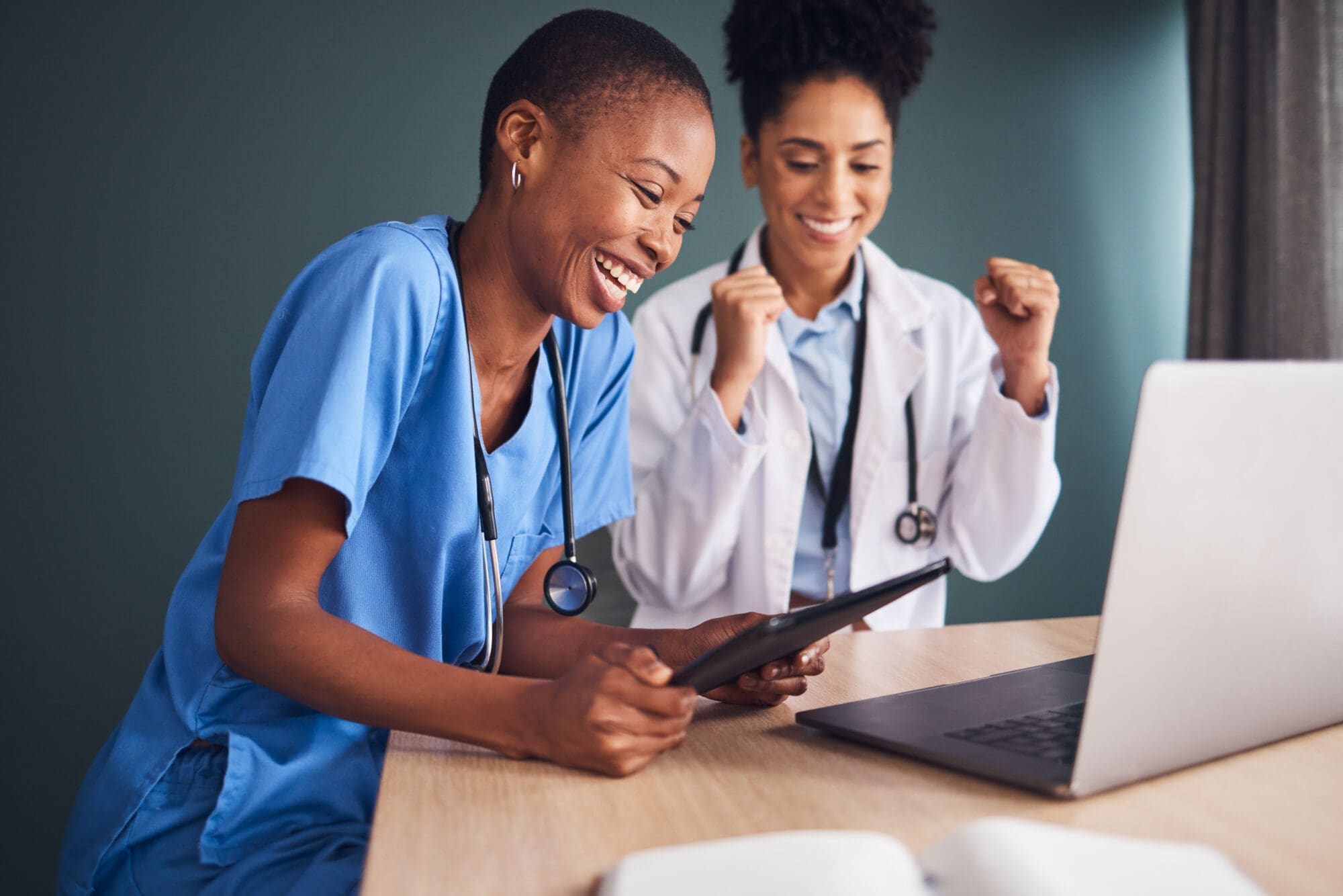 Two healthcare workers celebrate something on a laptop and tablet.